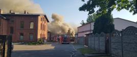 Zdjęcie przedstawia 2 samochody pożarnicze na tle budynku magazynowego z którego wydobywają się kłęby dymu.