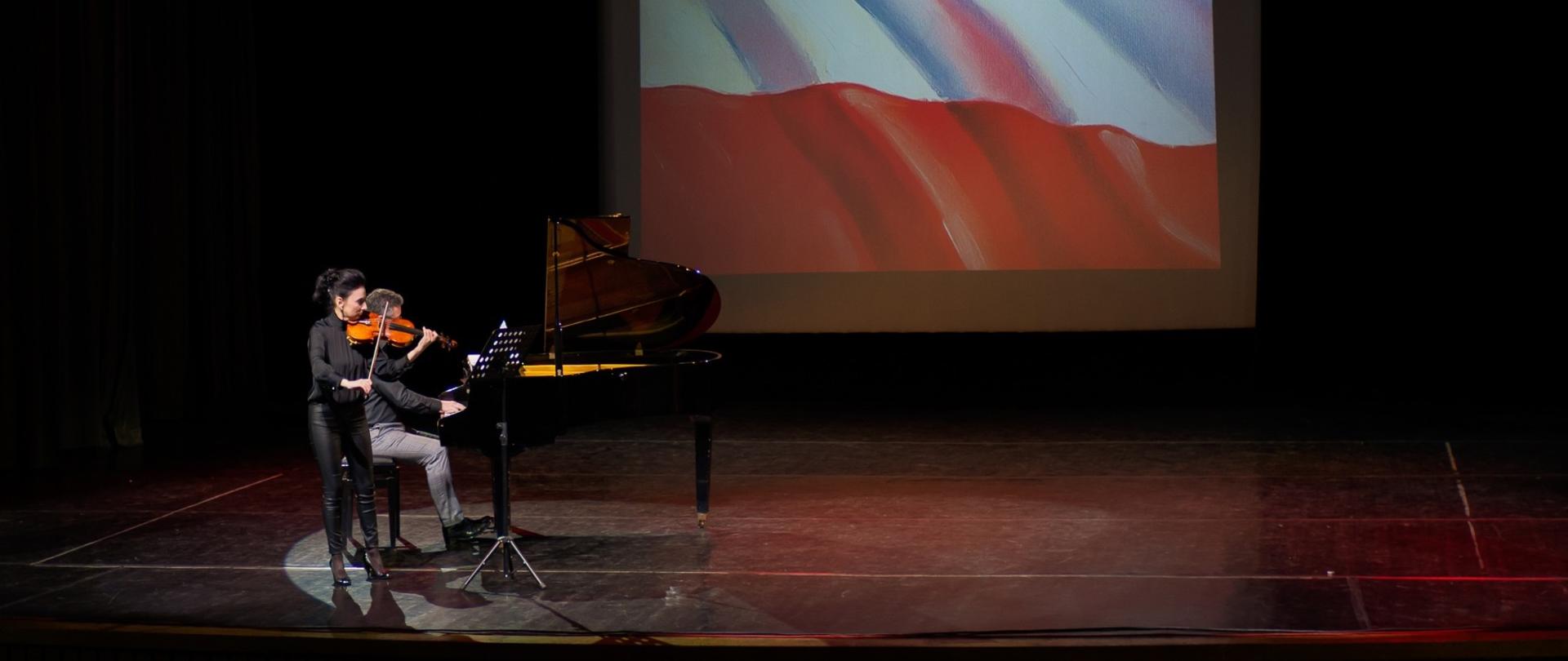 Koncert na scenie. Na czarnej, oświetlonej scenie, w ciemnym otoczeniu, dwoje artystów wykonuje utwór. Kobieta na pierwszym planie gra na skrzypcach, ubrana jest na czarno, w spodnie i szpilki. Siedzący obok niej mężczyzna gra na czarnym fortepianie. W tle, na dużym ekranie, widoczna jest projekcja malarska stylizowana na flagę Polski (czerwień i biel). Jest to ujęcie z Koncertu Patriotycznego.