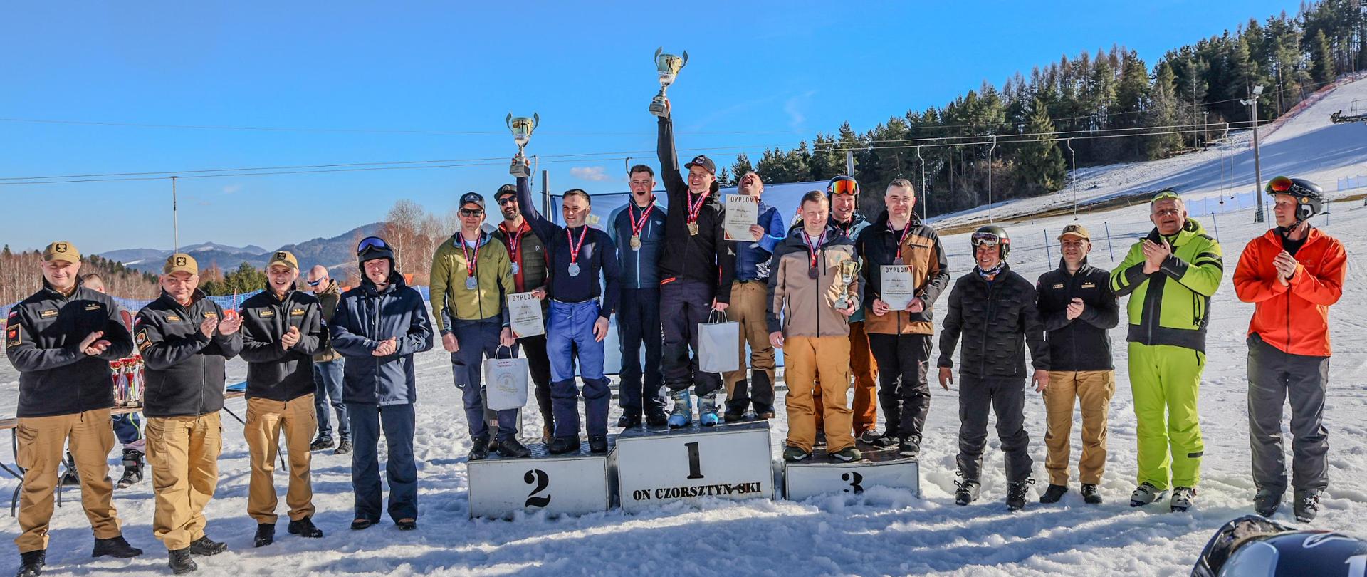 Ustawione u podnóża stoku na śniegu podium, na nim dziewięciu zawodników w sportowych strojach, po prawej i lewej stronie podium kilka osób pozujących do pamiątkowego zdjęcia. 