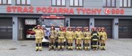 Przed garażem Jednostki Ratowniczo-Gaśniczej strażacy ubrani w ubrania specjalne stojąc w szeregu oddają hołd, w tle samochód ratowniczo - gaśniczy