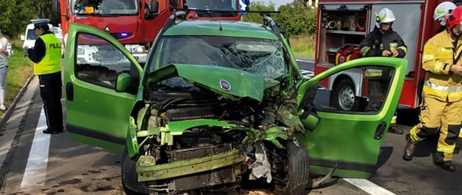 Na zdjęciu znajduje się rozbity samochód osobowy marki Fiat, kolor jasnozielony. Stoi na jezdni. Obok osoby postronne, strażacy i policjanci pracujący na miejscu zdarzenia.