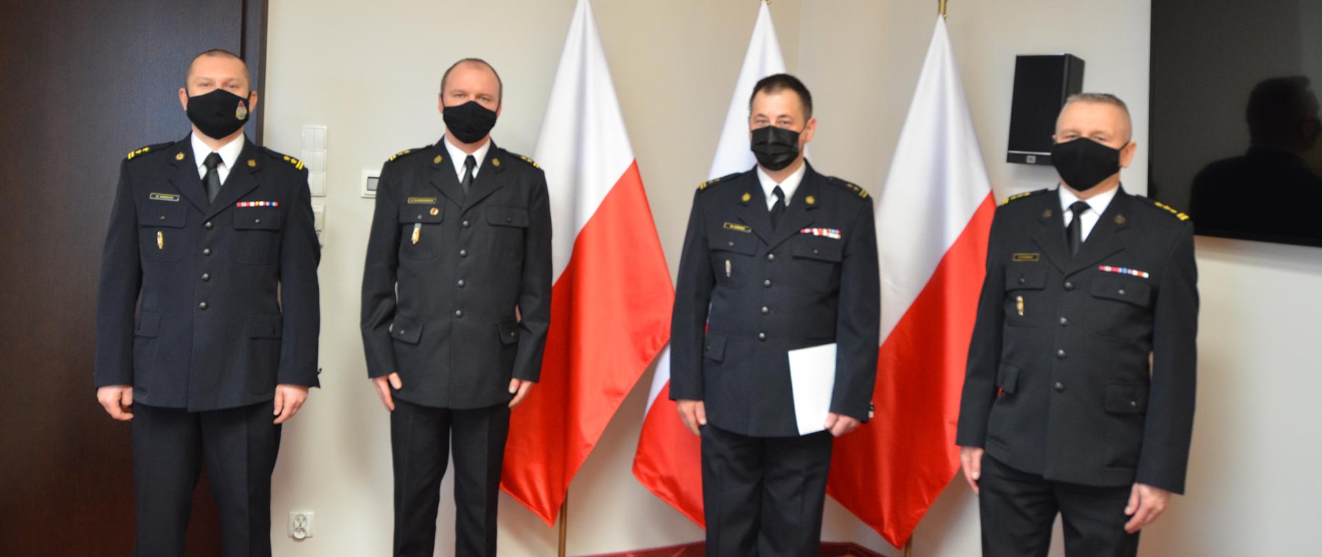 Na zdjęciu widać trzech strażaków w umundurowaniu wyjściowym. W tle trzy flagi Polski.