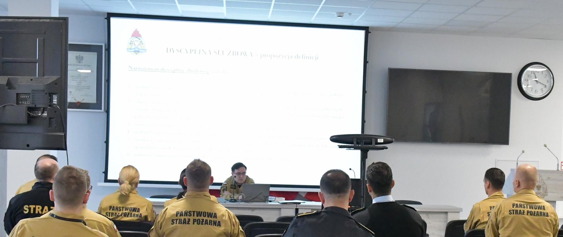 Strażacy w mundurach koloru pisakowego siedzą na krzesłach przed nimi za stołem siedzi funkcjonariusz Państwowej Straży Pożarnej siedzi przed laptopem i wykłada za nim na ekranie wyświetlane są tematy szkolenia obok znajduje się telewizor oraz zegar.