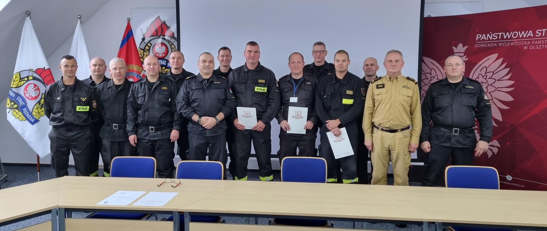 Na zdjęciu widać strażaków, instruktorów oraz komisję egzaminacyjną pozujących do zdjęcia grupowego w sali konferencyjnej. 