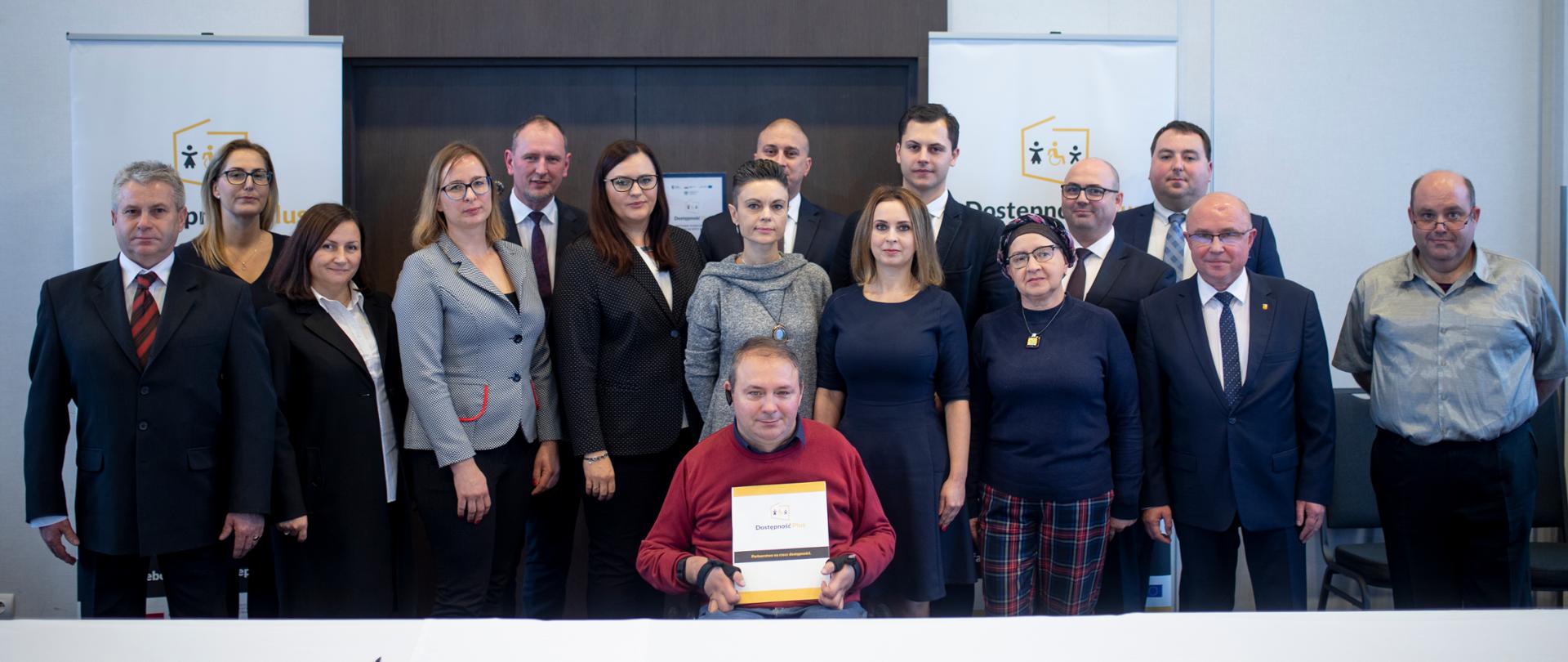 Szesnaście osób w dwóch rzędach, na środku jedna osoba na wózku inwalidzkim z porozumieniem w rękach. Z tyłu roll-upy z logo programu Dostępność Plus.