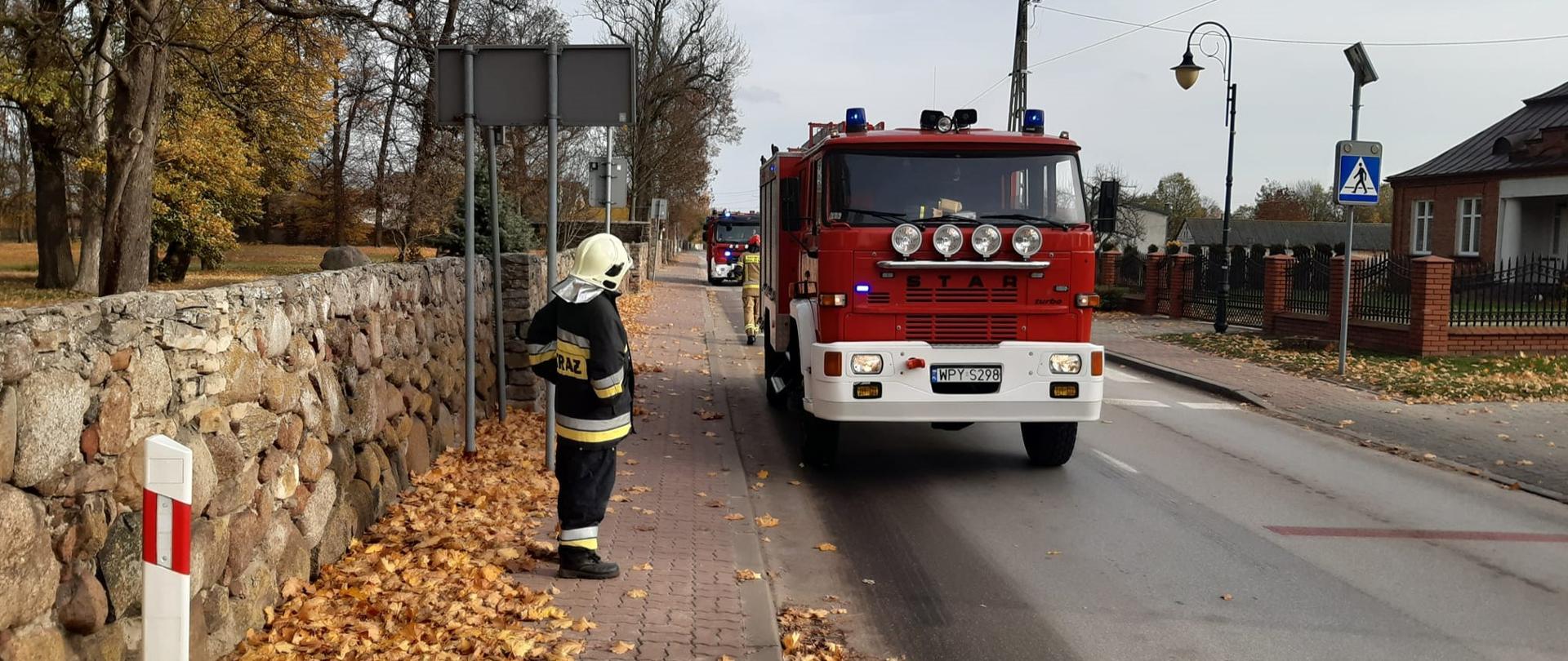 Zdjęcie przedstawia strażaka w pełnym umundurowaniu i hełmie, który stoi na chodniku wyłożonym kostką, obok kamiennego muru. Na chodniku leżą jesienne liście. Na ulicy widoczne są dwa czerwone wozy strażackie z włączonymi światłami błyskowymi. W tle widać budynki mieszkalne i drzewa z resztkami liści.