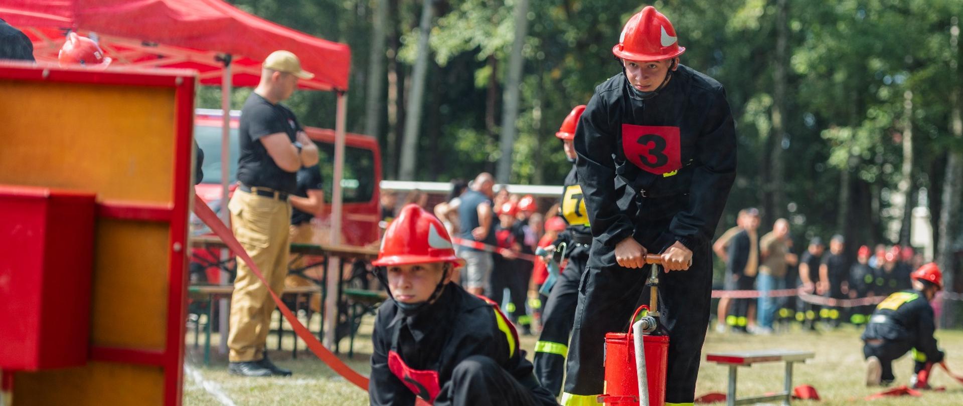 Na zdjęciu widoczne są zawody sportowo-pożarnicze młodzieżowych drużyn pożarniczych. Dwóch uczestników w czarnych ubraniach ochronnych z czerwonymi numerami startowymi i czerwonymi hełmami wykonuje zadanie bojowe. Jeden z nich klęczy przy prądownicy wodnej skierowanej w stronę celu, drugi obsługuje hydronetkę ręczną. W tle znajdują się namioty, samochód oraz osoby obserwujące zawody. Całość odbywa się na otwartej przestrzeni trawiastej w otoczeniu drzew.