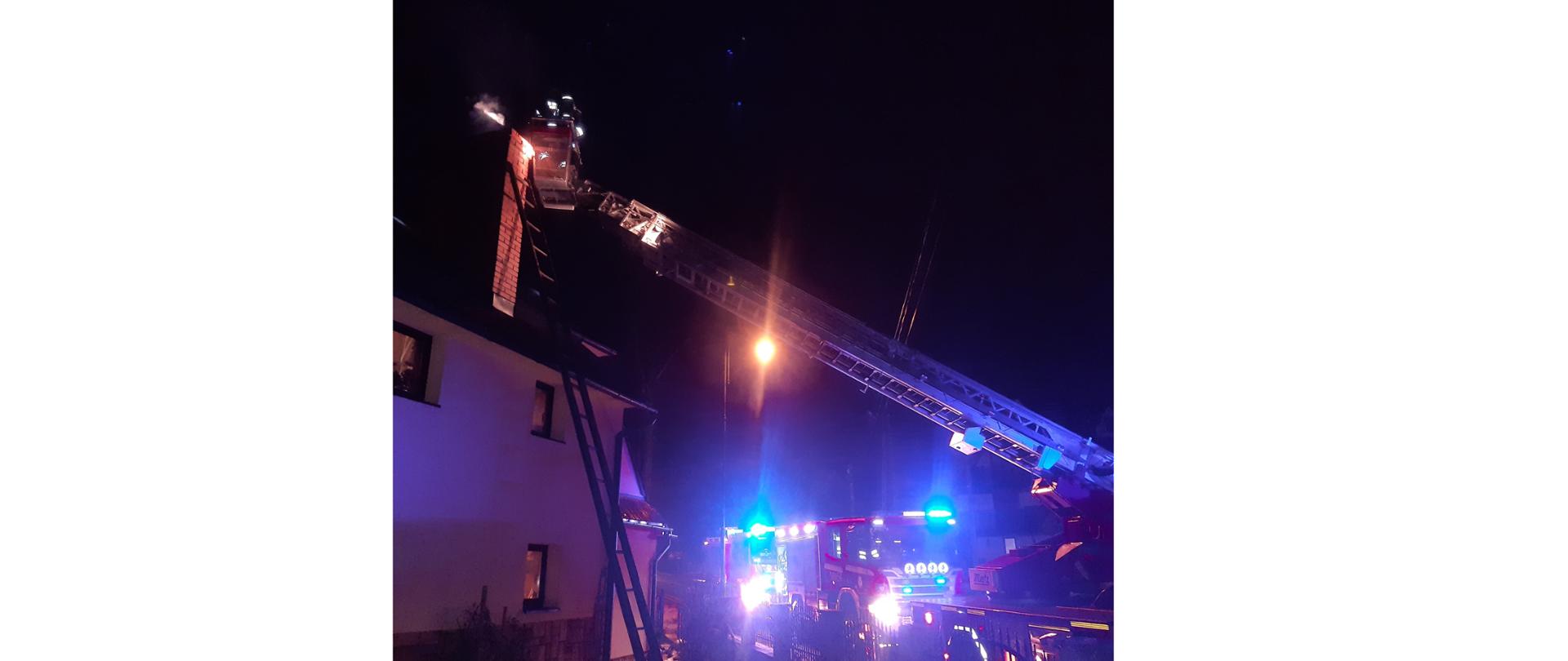 Zdjęcie przedstawia strażaków walczących z pożarem sadzy w miejscowości Białka Tatrzańska. Na zdjęciu strażacy w koszu drabiny mechanicznej, w tle wozy strażackie i ciemne tło, z uwagi na późne godziny prowadzonej akcji.