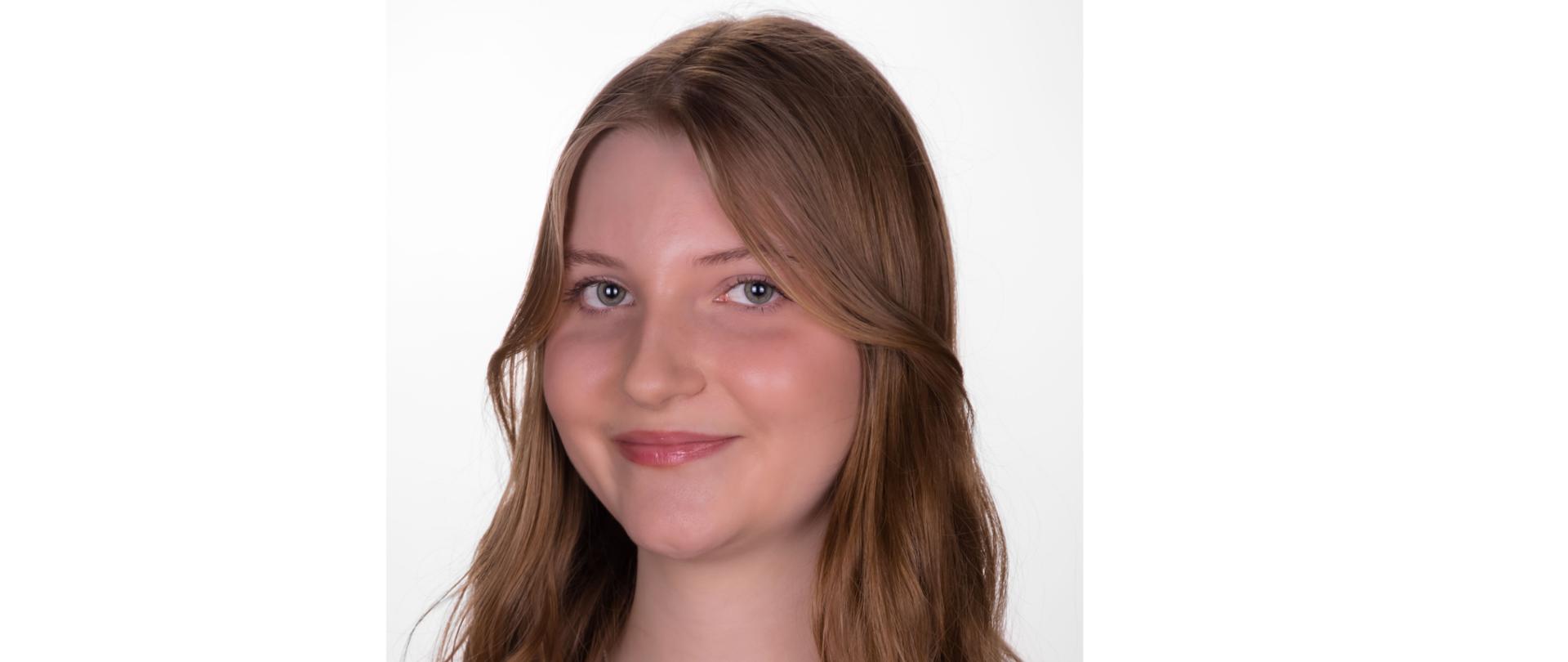 Zdjęcie kolorowe. Portret uśmiechniętej kobiety o jasnej karnacji, zielonych oczach i długich, pofalowanych włosach w kolorze jasnego brązu z blond refleksami. Kobieta ma delikatny makijaż, lekko błyszczące usta i delikatny łańcuszek na szyi. Ubrana w ciemnobordową koszulkę polo z rozpiętymi guzikami. Tło jednolite, białe.