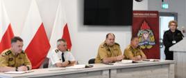 Kierownictwo Komendy Wojewódzkiej Państwowej Straży Pożarnej w Gdańsku siedzi za zostałem na którym leżą kalendarze. Jeden z funkcjonariuszy w umundurowaniu w kolorze czarnym stoi za mównicą. 