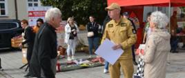 Strażak omawia alerty RCB oraz znaczenie sygnałów alarmowych trzyma plakat edukacyjny „Poznaj Alerty RCB o zagrożeniu atakiem z powietrza” opisujący trzy poziomy alarmowe (Czujność, Gotowość, Alarm) oraz zalecenia dotyczące zachowania w budynku i na zewnątrz.