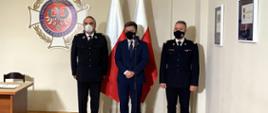 na zdjęciu widać komendanta głównego PSP nadbrygadiera Andrzej Bartkowiaka w umundurowaniu galowym, druha Waldemara Pawlaka prezesa zarządu głównego związku ochotniczych straży pożarnych rzeczpospolitej polskiej, Michała Dworczyka, szefa Kancelarii Prezesa Rady Ministrów w garniturze. na ścianie widać logo związku ochotniczych straży pożarnych RP, za stojącymi są dwie flagi polski.