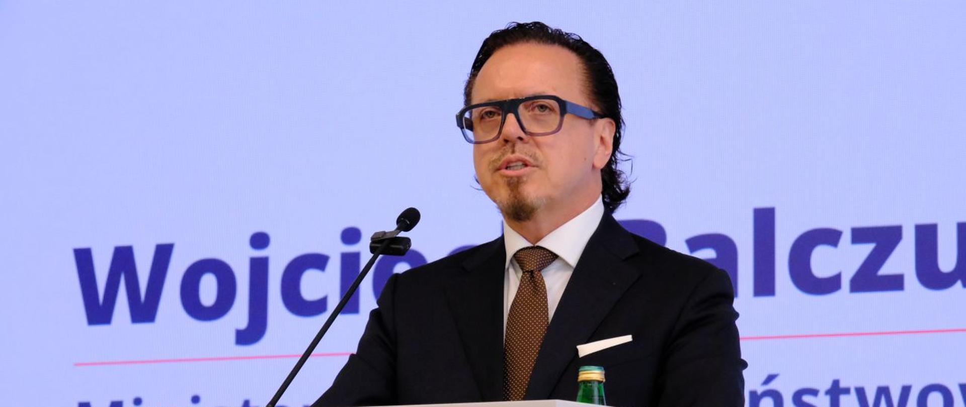 Minister Wojciech Balczun stoi za mównicą. W tle ekran. 