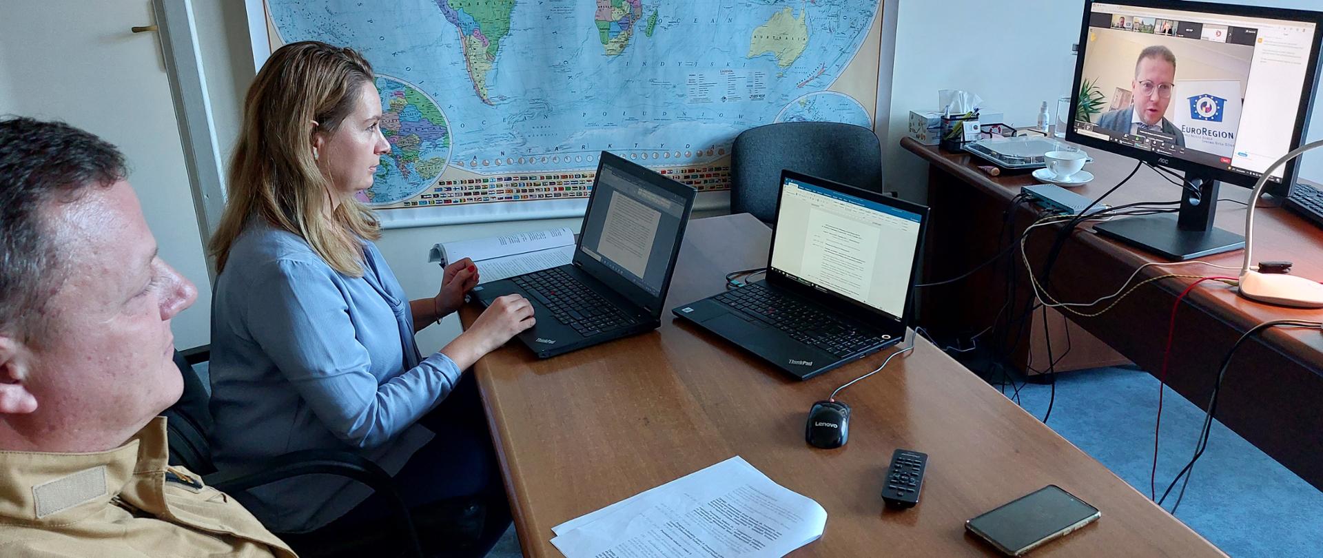 Dwie osoby siedzą przy biurku przy monitorach ekranowych uczestnicząc w wideokonferencji