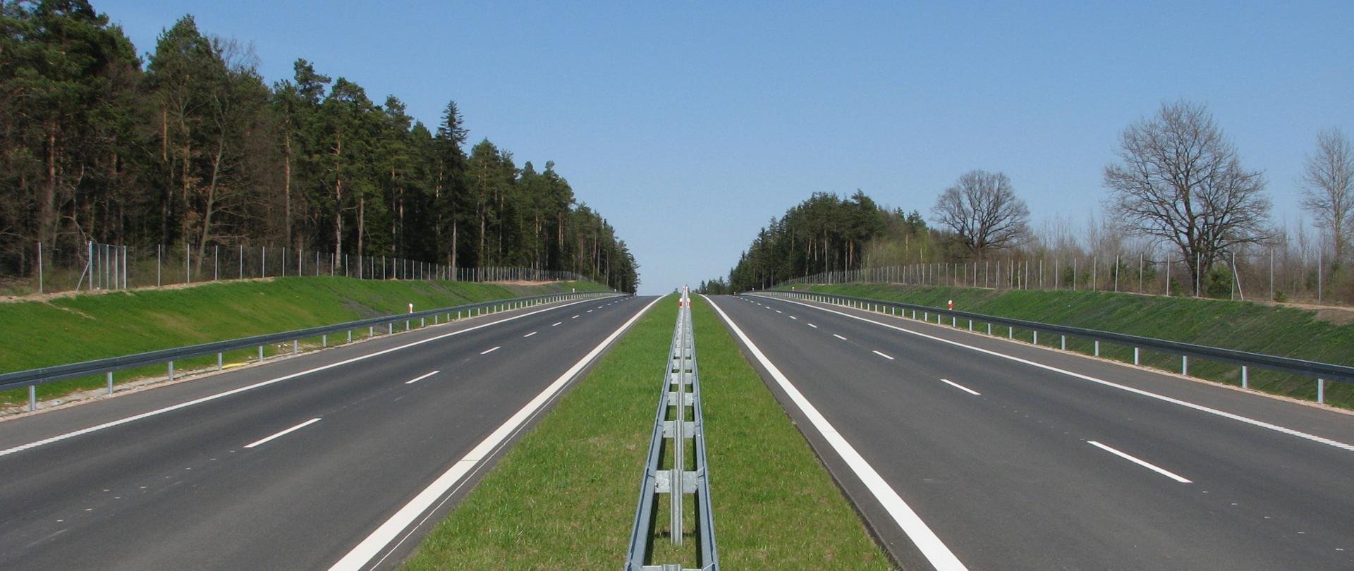Droga ekspresowa - dwie jezdnie przedzielone pasem zieleni i barierą, przy drodze za ogrodzeniem las
