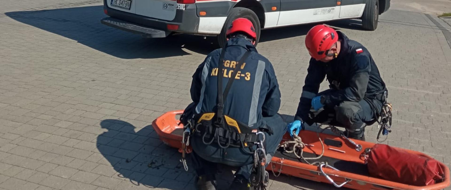 Zdjęcie przedstawia dwóch ratowników pochylonych nad noszami wannowymi sprawdzając i układając liny po ich użyciu. Za nimi stoi ambulans Państwowego Pogotowia Ratowniczego.