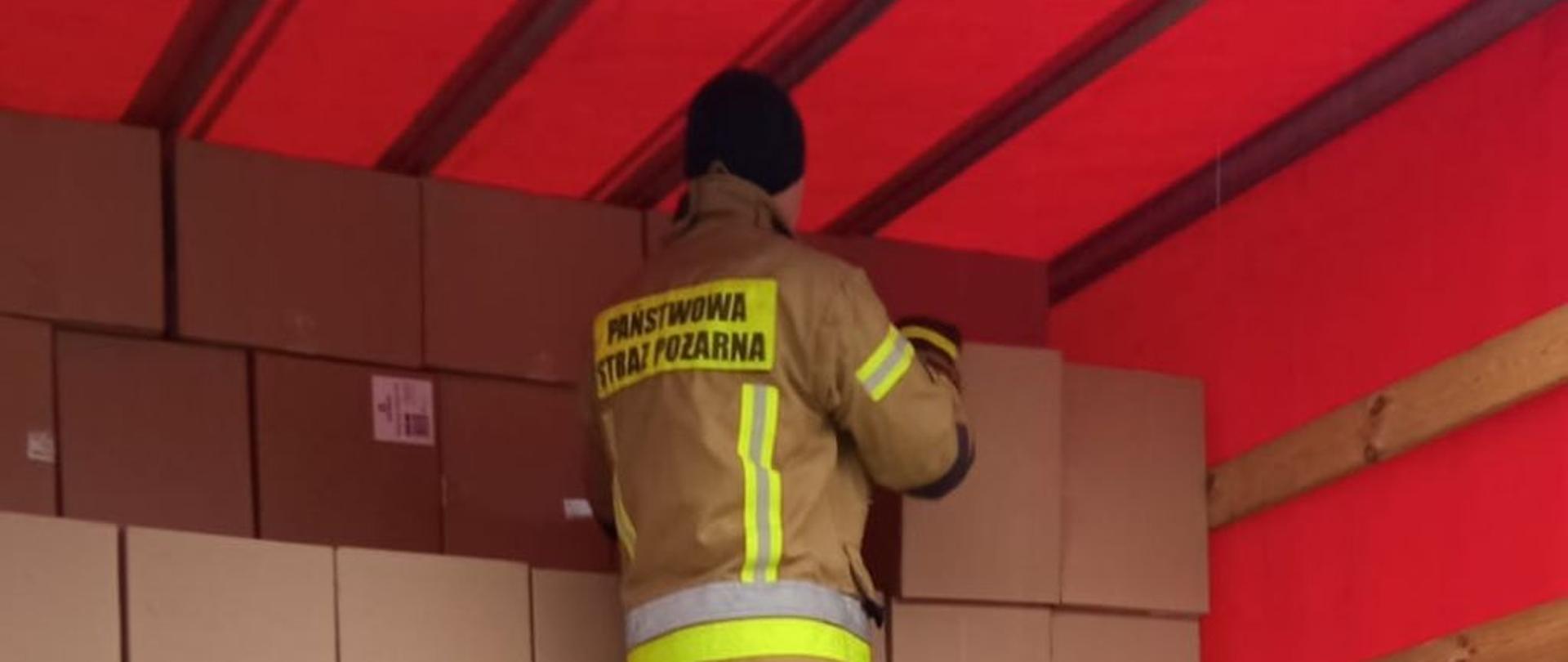 Jeden ze strażaków ubrany w piaskowy mundur bojowy podaje brązowe kartony z części ładunkowej samochodu ciężarowego. Drugi strażak stojący ma ziemi ubrany w czarny mundur bojowy odbiera kartony.