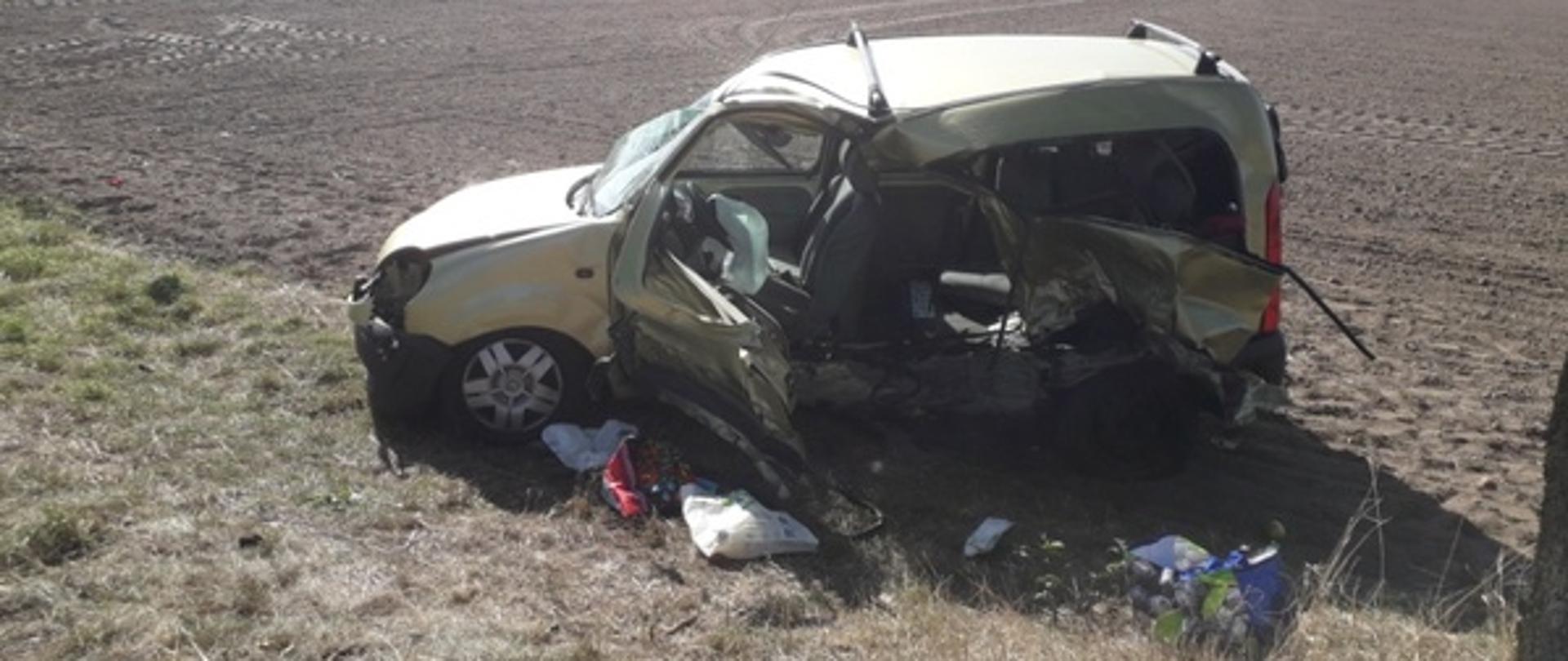 Słonecznie. Żółte zniszczone auto osobowe stojące częściowo na trawie pobocza i zaoranym polu uprawnym. Pojazd z bardzo dużymi uszkodzeniami po stronie kierowcy. Na zewnątrz pojazdu leżą torby zakupowe oraz porozrzucane sprawunki. W oddali zielone pole i zabudowania oraz drzewa.