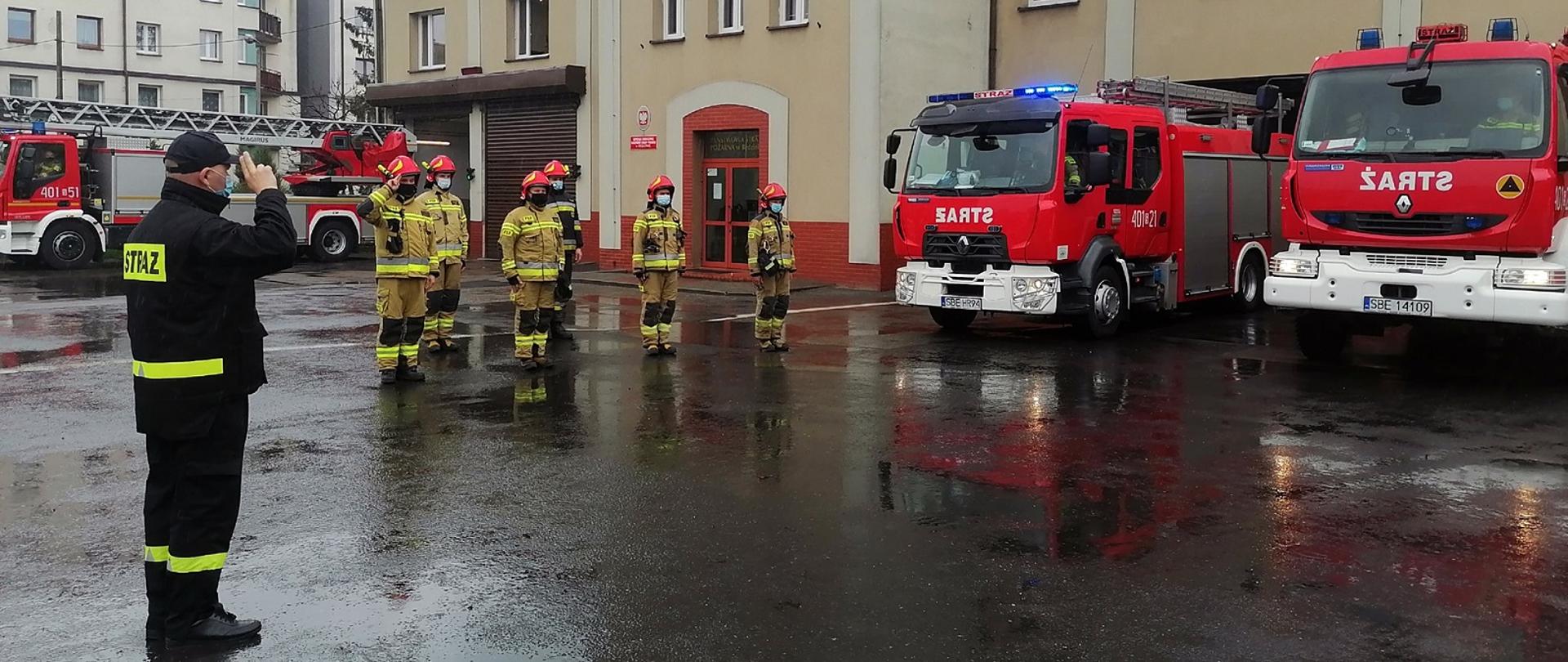Zdjęcie przedstawia strażaków oddających hołd Policjantowi poległemu na służbie. Zdjęcie wykonano na placu przed budynkiem Komendy Powiatowej Państwowej Straży Pożarnej w Będzinie. Przed budynkiem stoją trzy samochody pożarnicze.