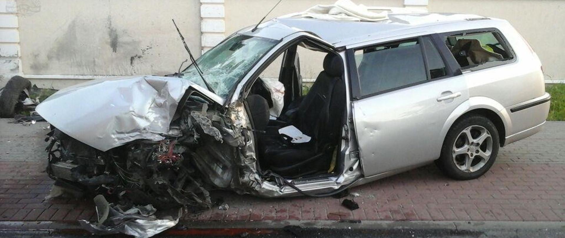 Rozbity srebrny samochód na tle ogrodzenia. Auto znajduje się na chodniku.