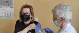 Zdjęcie przedstawia druhnę Katarzynę Czerwińską z OSP Lisewo w momencie
szczepienia przez pielęgniarkę w punkcie szczepień w szpitalu w Chełmnie. Druhna siedzi
na kozetce, siedząc obok niej pielęgniarka wykonuje szczepienie w lewe ramie.