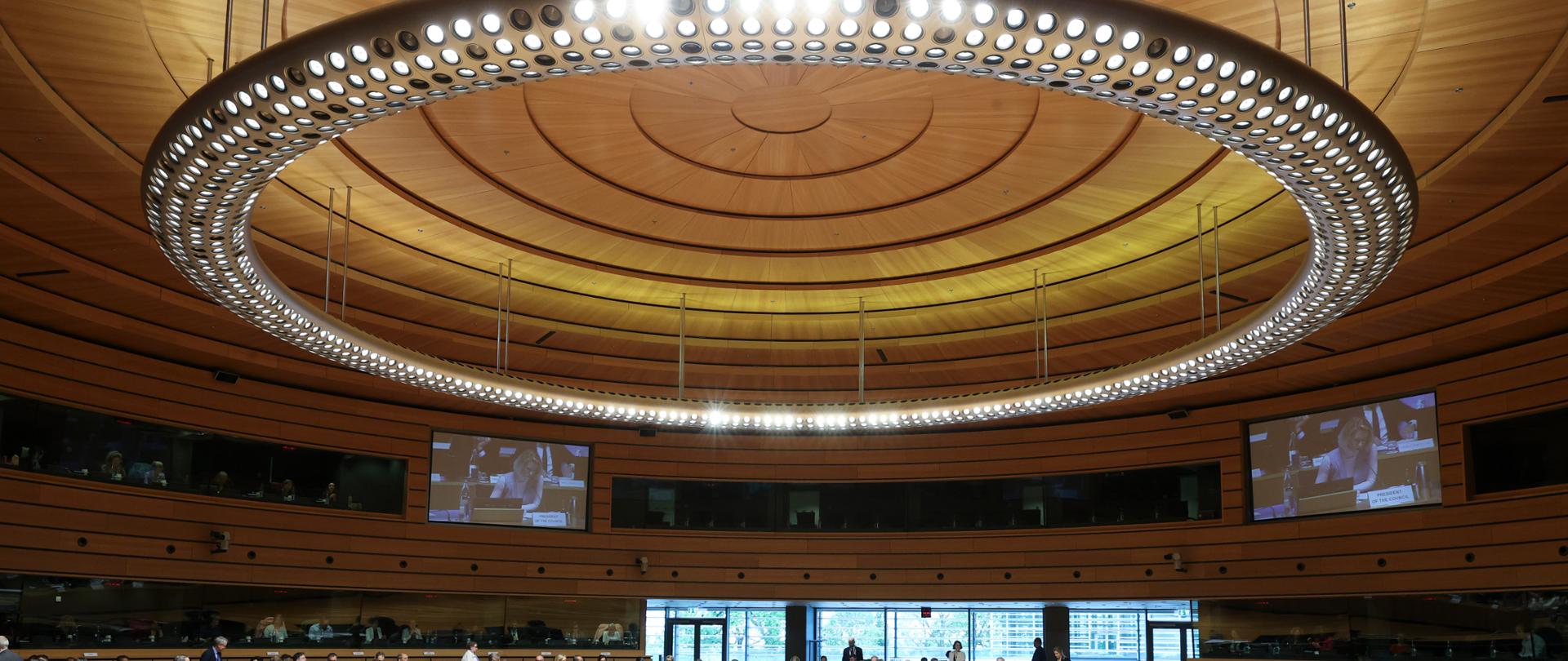 Many people sitting around circular table. Above them huge wooden celiling with many flight, circular lights.