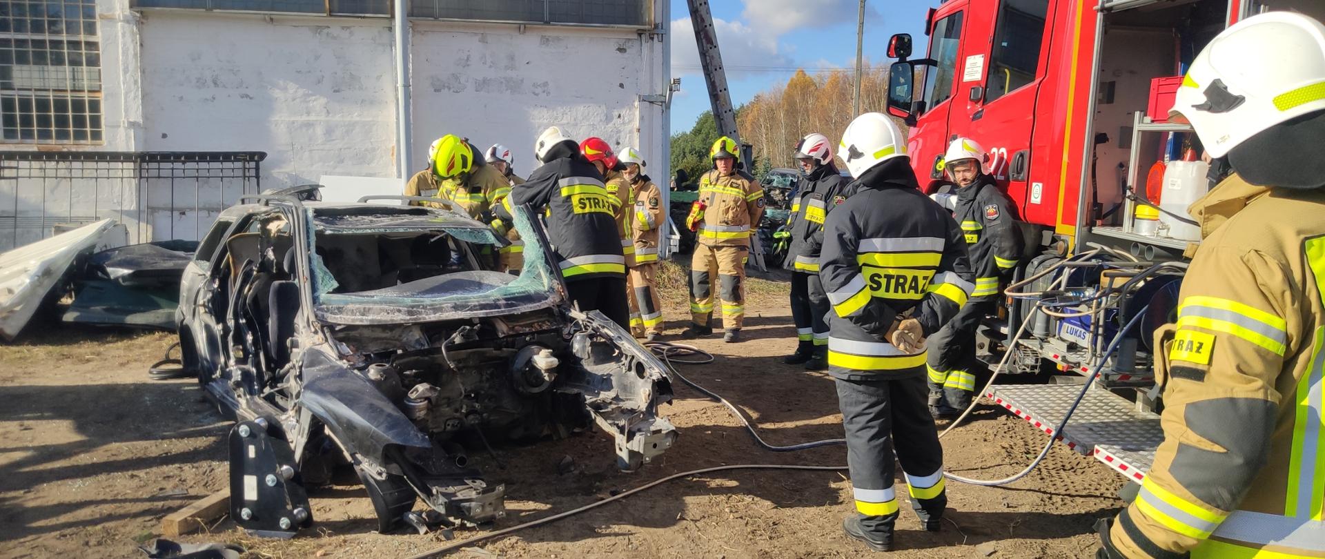 Zdjęcie przedstawia teren złomowiska pojazdów – plac ćwiczeń. Na placu znajduje się wrak samochodu osobowego oraz średni samochód ratowniczo gaśniczy w którym znajduje się pompa hydrauliczna zasilająca za pomocą węży urządzenia. Strażacy ochotniczych straży pożarnych pod okiem instruktora ćwiczą usuwanie za pomocą urządzeń hydraulicznych boku pojazdu celem wykonania dostępu do osoby poszkodowanej.
