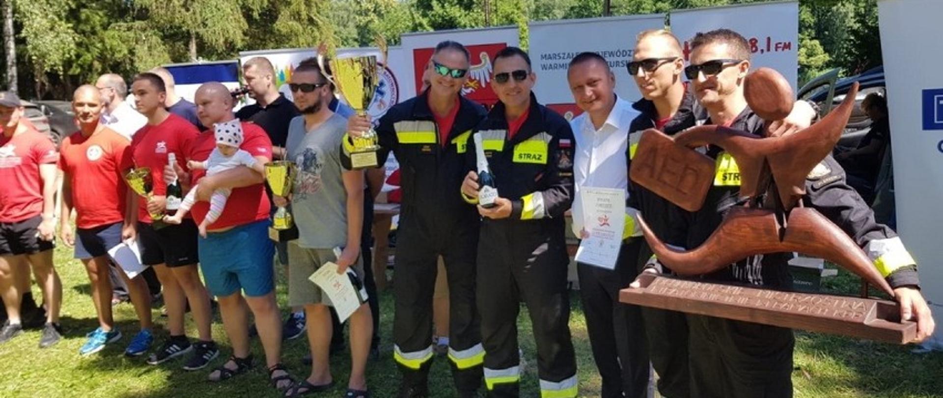 Grupa piętnastu mężczyzn stoi w szeregu, wśród nich jest czterech ubranych w mundury strażackie jeden z nich trzyma drewnianą statuetkę, a kolejny trzyma puchar