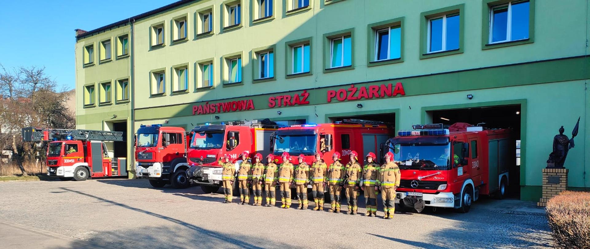Budynek KM PSP w Piotrkowie Trybunalskim. Strażacy w szeregu na tle samochodów ratowniczych.
