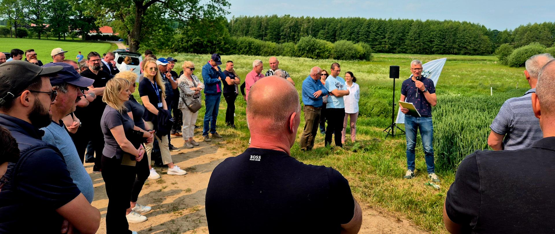 Mężczyzna stoi na polu w granatowej koszulce i trzyma mikrofon. Tłum ludzi stoi zwrócony w stronę mężczyzny z mikrofonem. 