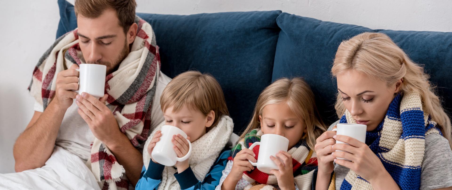 Na zdjęciu jest rodzina. Mama, tata oraz syn i córka. Leżą w łóżku z herbatą, owinięci szalikami.