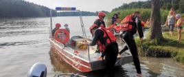 Na zdjęciu ratownicy zabezpieczeni w łodzi ratowniczej na brzegu oraz ludzie będący uczestnikami pokazów w tle jezioro