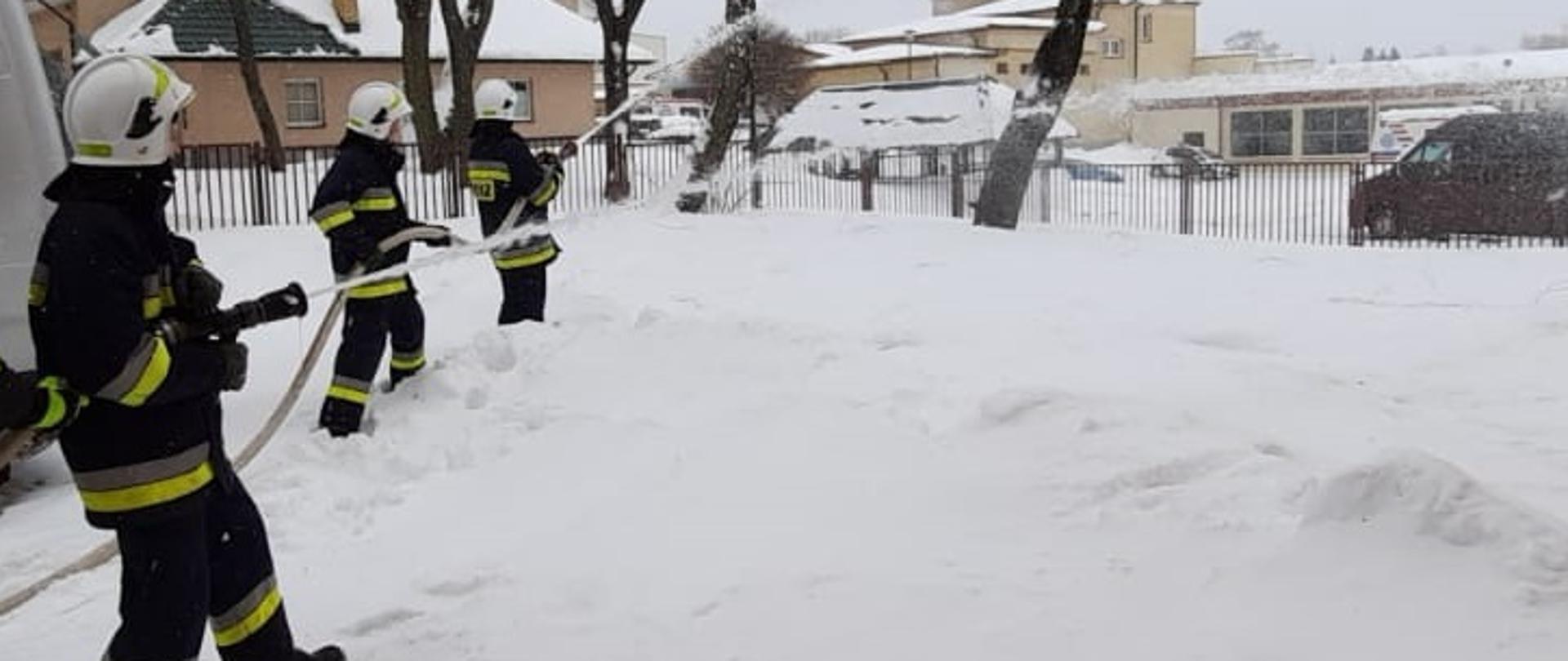 Zdjęcie obrazuje 3 strażaków trzymających węże z których leci woda na ziemi leży śnieg w tle widać drzewa i budynki