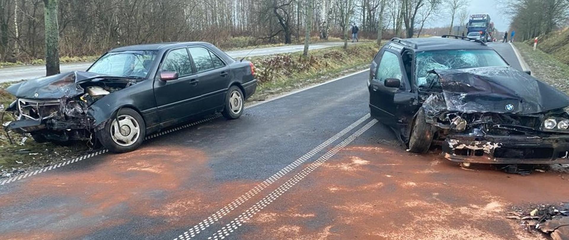 Dwa czarne rozbite samochody osobowe stoją na drodze. Mają Uszkodzone przody. Za nimi w oddali widać samochody samochody stojące w korku, który powstał w wyniku zablokowania drogi przez rozbite pojazdy.Widoczni też są w oddali ludzi, którzy przyglądają się zdarzeniu. 