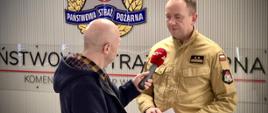 Strażak udziela wywiadu mężczyźnie, który trzyma przed nim mikrofon. W tle logo PSP. Strażak w ręku trzyma kartkę z nadrukiem.