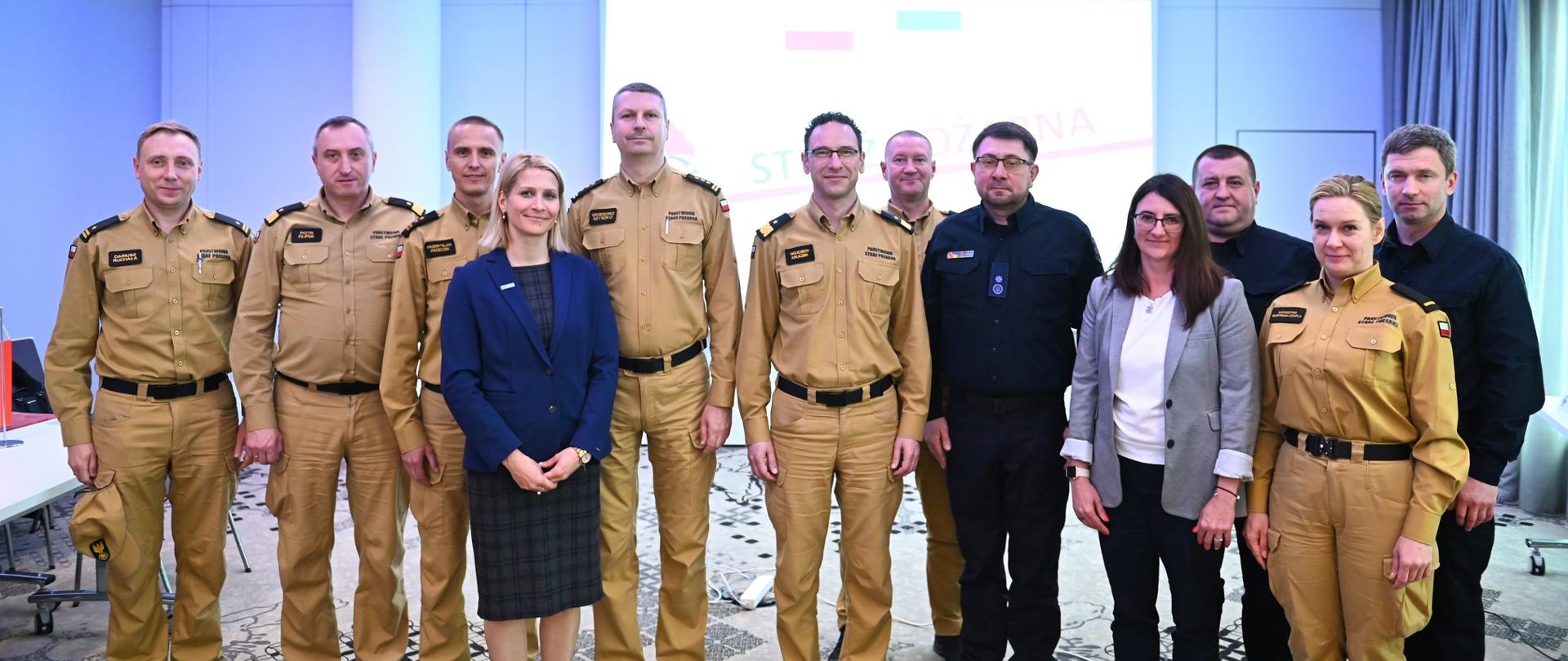 Zdjęcie grupowe przedstawicieli PSP w służbowych mundurach koloru żółtego, ukraińskich służb ratowniczych w czarnych mundurach oraz osób cywilnych, wykonane po zakończeniu spotkania na tle ekranu z logo PSP i flagami Polski i Ukrainy.
