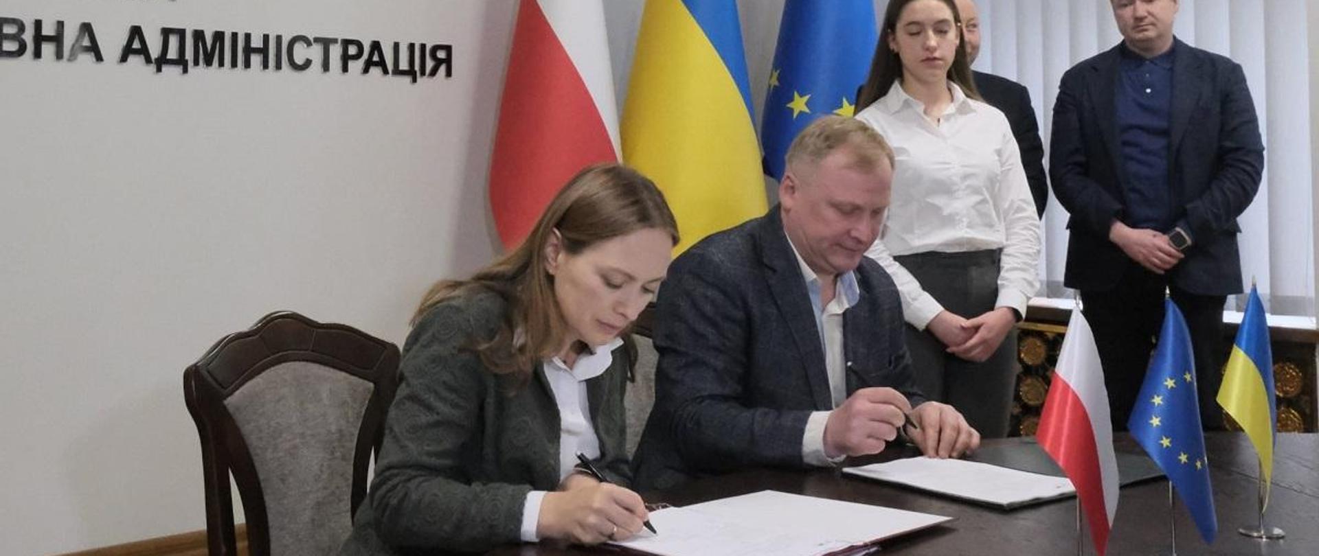 Minister funduszy i polityki regionalnej Katarzyna Pełczyńska-Nałęcz siedzi przy stole i podpisuje dokument, obok niej po prawej stronie siedzi mężczyzna