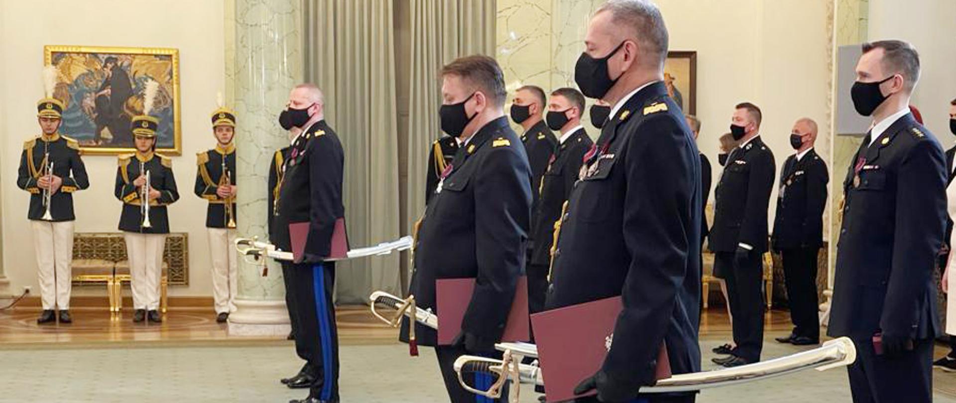 strażacy stojący w umundurowaniach galowych trzymający szable generalskie , w tle osoby towarzyszące
