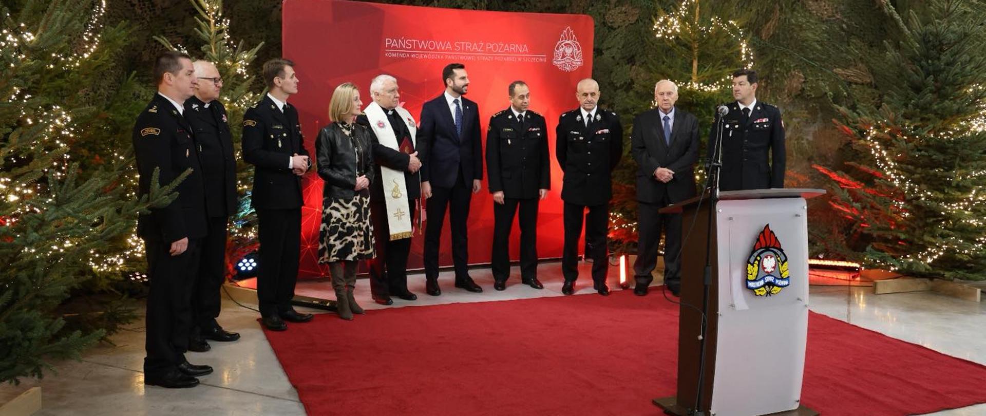 a uroczystej scenie stoi grupa osób w mundurach strażackich i strojach formalnych. Jedna osoba przemawia przy mównicy. Tło stanowią choinki, świąteczne oświetlenie oraz czerwony baner Państwowej Straży Pożarnej. Atmosfera jest oficjalna i uroczysta.