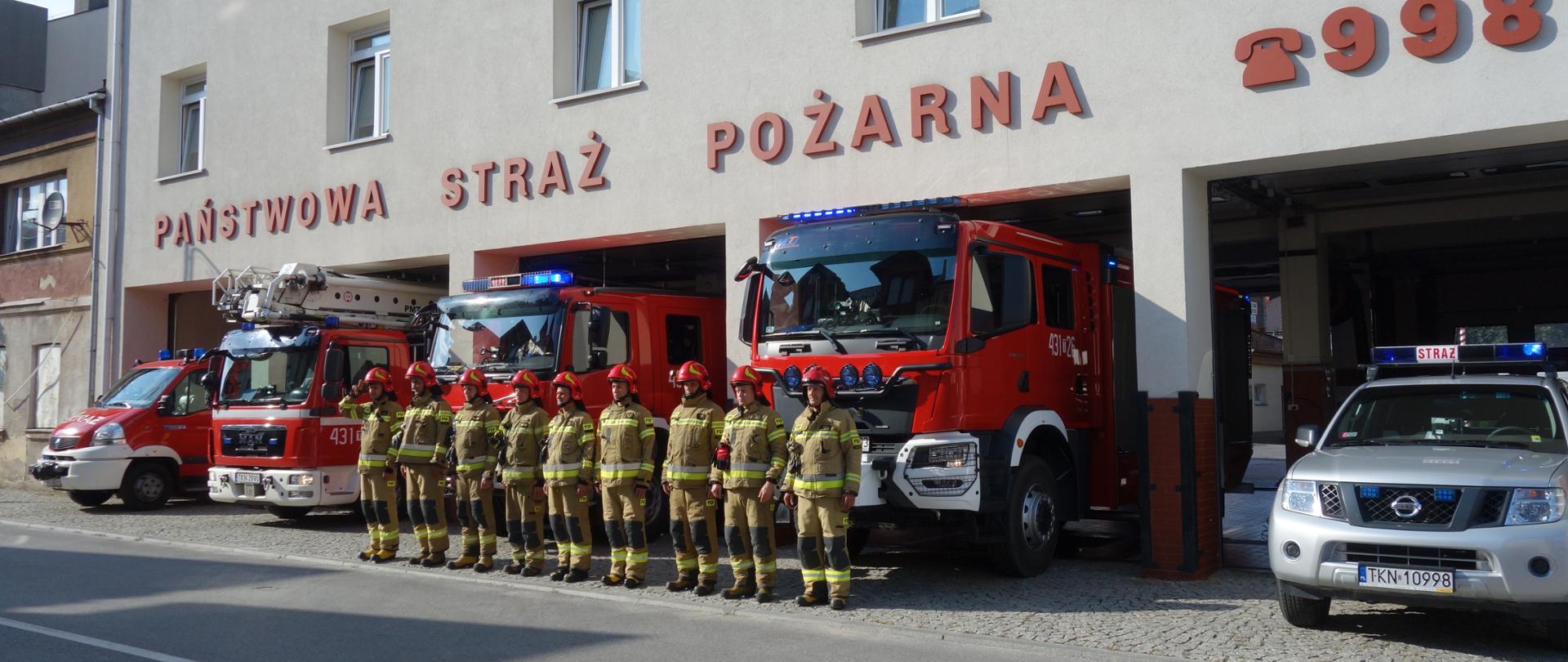 Zbiórka koneckich strażaków z okazji 78 rocznicy Powstania Warszawskiego. Przed garażem stoją pojazdy pożarnicze a przed nimi w szeregu strażacy w ubraniach bojowych.