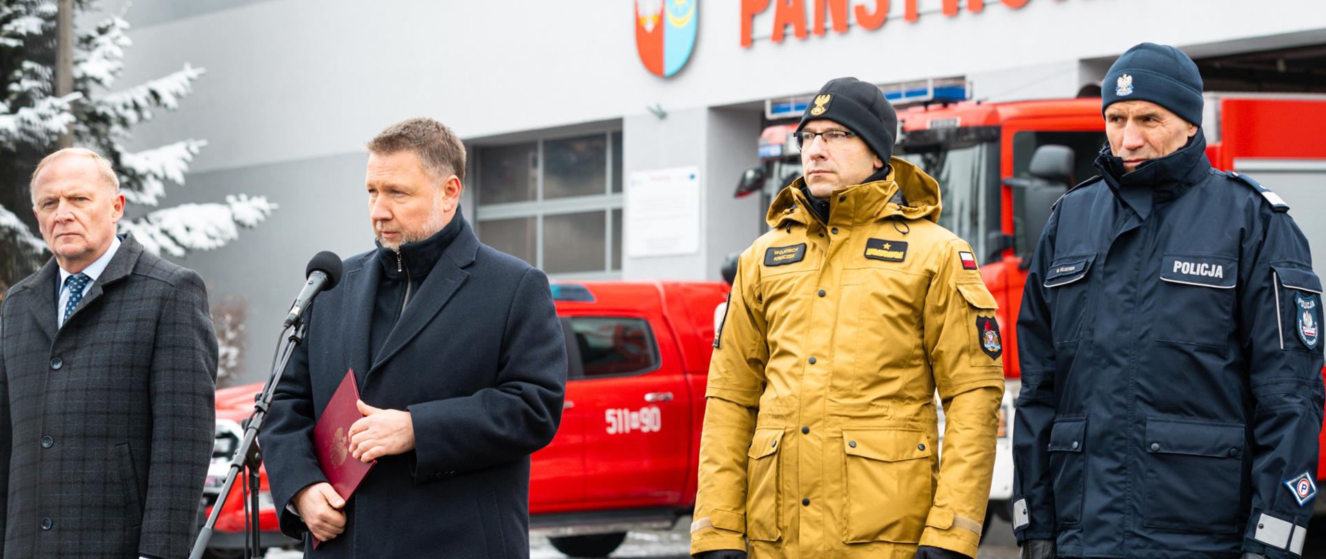 Czterech mężczyzn stoi przed budynkiem Państwowej Straży Pożarnej; dwóch w eleganckich płaszczach, jeden w mundurze PSP, jeden w mundurze Policji. Mężczyzna na środku przemawia do mikrofonu.