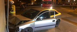 samochód osobowy uderzył w budynek przy drodze. Obok uszkodzonego samochodu stoi strażak a w oddali widać auto Straży Pożarnej. Miejsce zdarzenia odgrodzone jest biało-czerwoną taśmą, za którą stoją osoby postronne. 