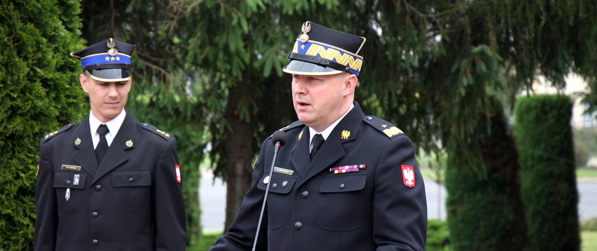 strażak w rogatywce i mundurze stoi przed mównicą, za nim strażak w rogatywce i mundurze z czerwona teczką w ręku