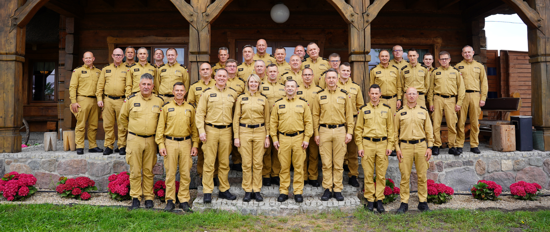 Na zdjęciu Pani i Panowie Komendanci stoją w grupie na schodach, na wejściu do budynku, uśmiechając się wspólnie - pozując do zdjęcia grupowego. Są ustawieni równo w szeregach. W tle budynek, przed budynkiem strażacy, obok nich czerwone kwiaty zasadzone równo w rzędzie po jednej i po drugiej stronie.