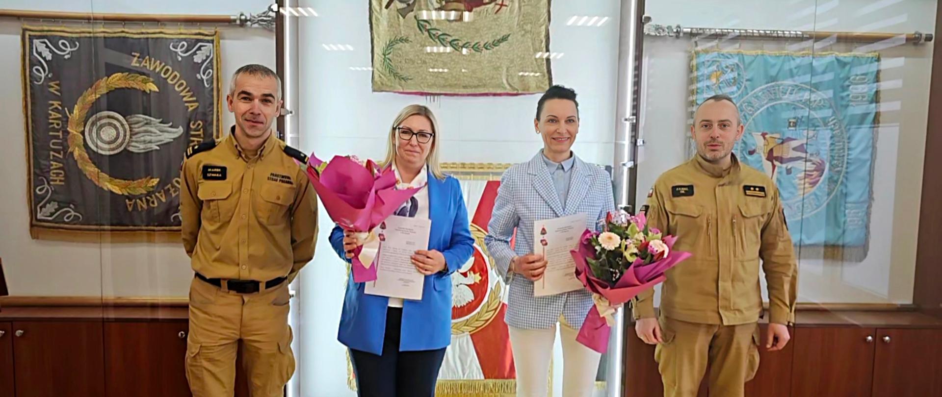 Zdjęcie przedstawia pracowniczki służby cywilnej pozujące do zdjęcia z Komendantami. Panie trzymają listy gratulacyjne oraz kwiaty. W tle gablota ze sztandarami oraz pucharami. 