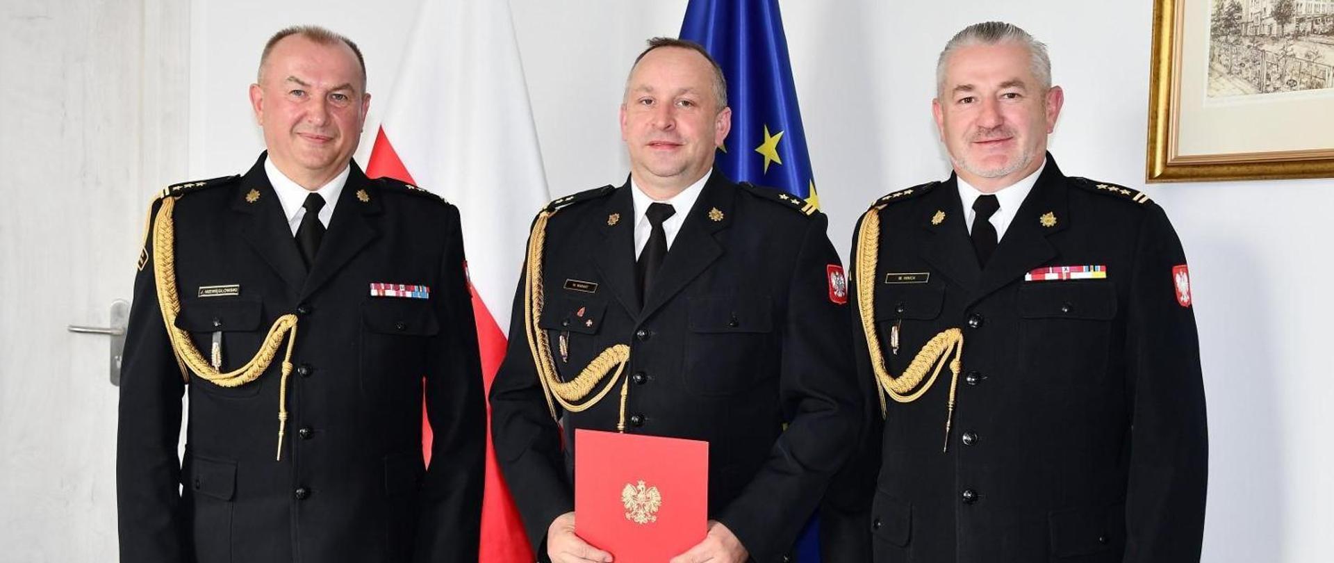 Na zdjęciu wiać trzech strażaków w mundurach wyjściowych koloru czarnego. Jeden trzyma czerwona teczkę akt powołania na stanowisko komendanta powiatowego 