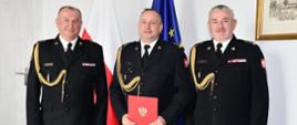 Na zdjęciu wiać trzech strażaków w mundurach wyjściowych koloru czarnego. Jeden trzyma czerwona teczkę akt powołania na stanowisko komendanta powiatowego 