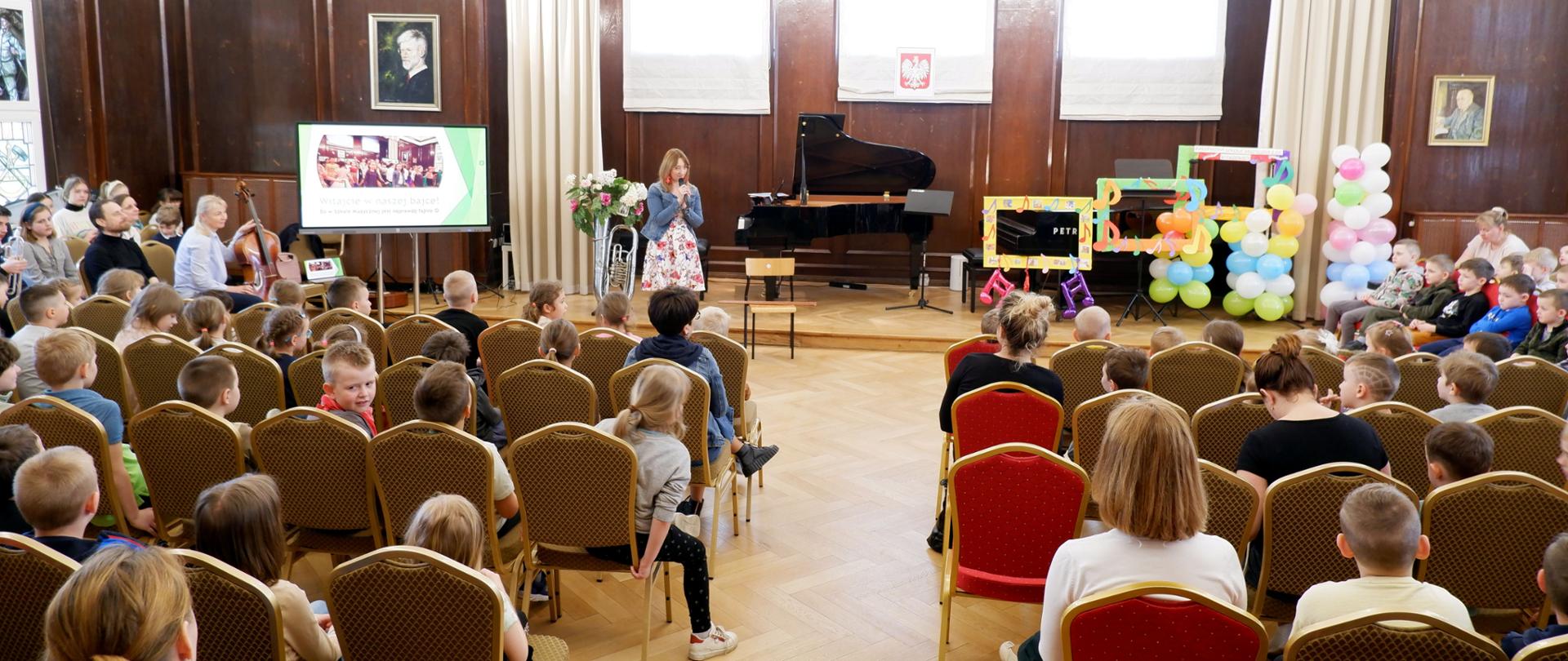Zdjęcie przestawia dzieci siedzące w auli szkoły muzycznej. Dzieci słuchają panią która na środku sceny opowiada o instrumentach, które wyświetlane są na ekranie telewizyjnym po lewej stronie sceny.