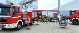 Na placu przed budynkiem przemysłowym ustawione są trzy wozy strażackie, w tym podnośnik hydrauliczny z koszem oraz samochody gaśnicze. W centrum kadru kilku strażaków w pełnym umundurowaniu PSP i OSP przygotowuje się do dalszych działań operacyjnych. Na dalszym planie, przed namiotem magazynowym, widoczna jest grupa obserwatorów – prawdopodobnie pracowników zakładu lub uczestników szkolenia. Na ziemi rozłożone są węże strażackie, a cała scena wygląda na element ćwiczeń ratowniczo-gaśniczych.