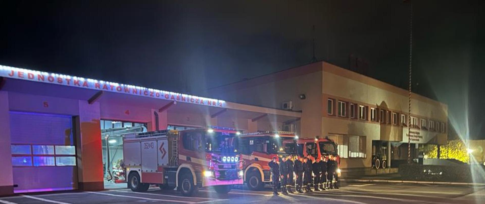 Strażacy Jednostki Ratowniczo-Gaśniczej numer 2 w Częstochowie stojąc w szeregu oddają hołd strażakowi, który zginął na służbie, w tle samochody strażackie z włączonymi niebieskimi sygnałami świetlnymi oraz budynek Jednostki Ratowniczo-Gaśniczej.